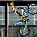 En motorcyklist i gul og sort dragt laver en wheelie på en trialcykel foran en industriel baggrund med vinduer og paller